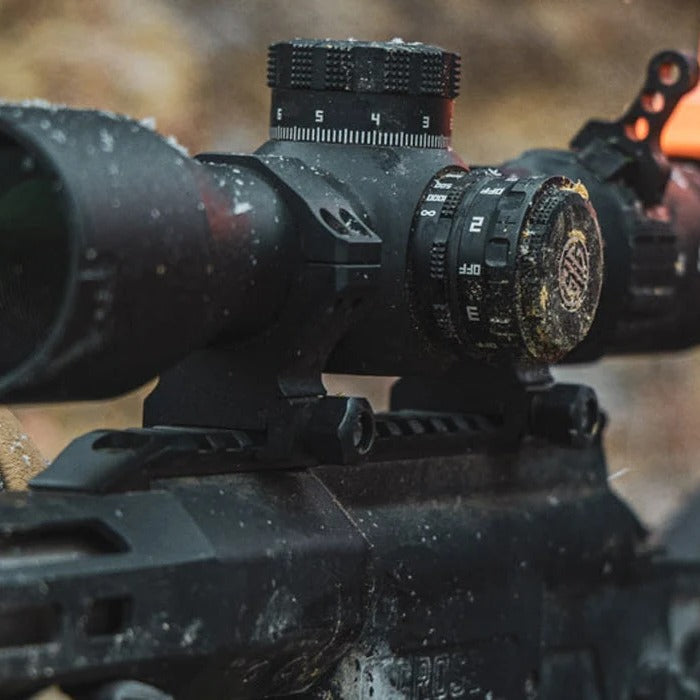 A close-up action shot of a hunter wearing an orange cap and protective face mask, aiming a black rifle with a mounted scope. The background is a blurry natural outdoor setting.
